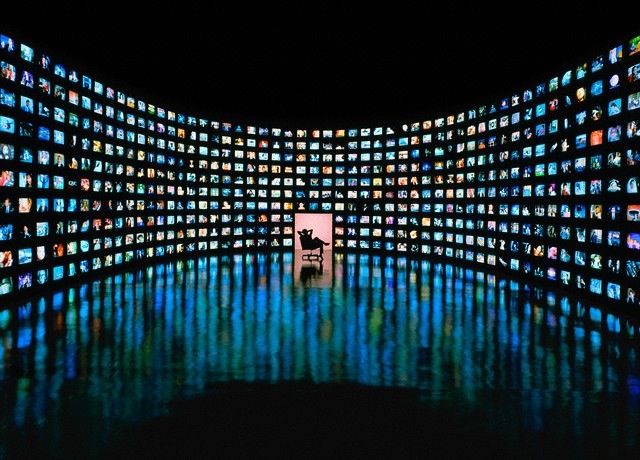 Curved wall of hundreds of illuminated screens surrounding a dark silhouette, evoking constant monitoring